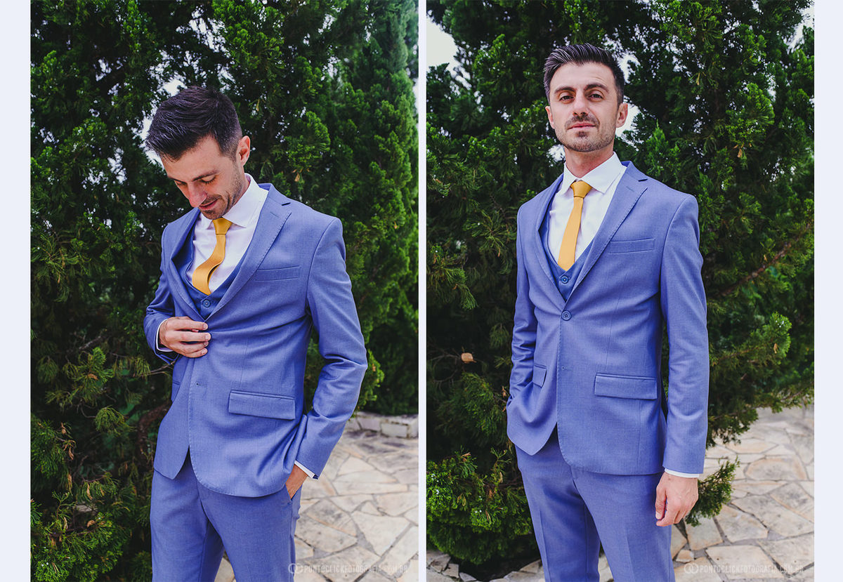 O noivo posa com um terno azul claro e uma gravata amarela, diante de um vegetação verde. Ele exibe um sorriso descontraído enquanto ajeita o paletó