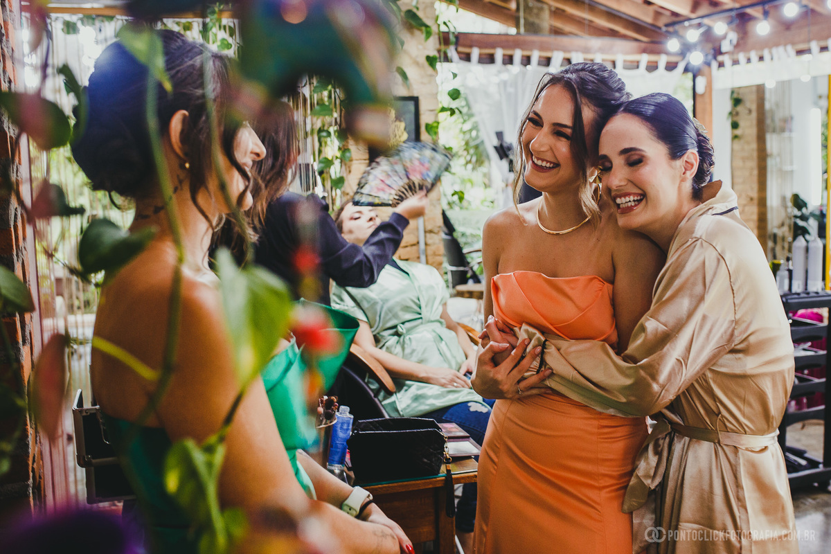 A noiva, em robe dourado, abraça uma madrinha de vestido laranja enquanto sorriem. Ao fundo, outra madrinha é atendida. Plantas pendentes e luz suave compõem um ambiente acolhedor, destacando a alegria e a cumplicidade do momento especial entre as amigas