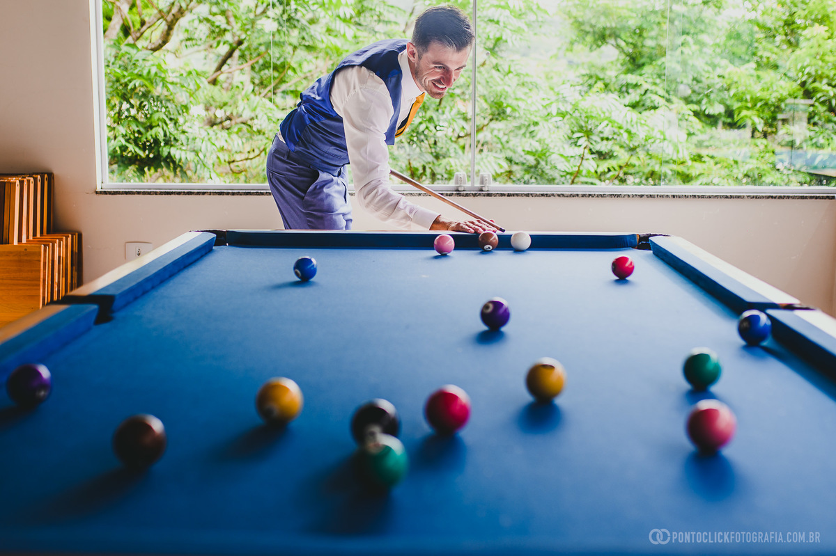 O noivo, com terno azul claro, joga bilhar com um sorriso largo, enquanto as bolas coloridas se alinham na mesa de sinuca. A luz natural entra pela janela, criando uma atmosfera tranquila