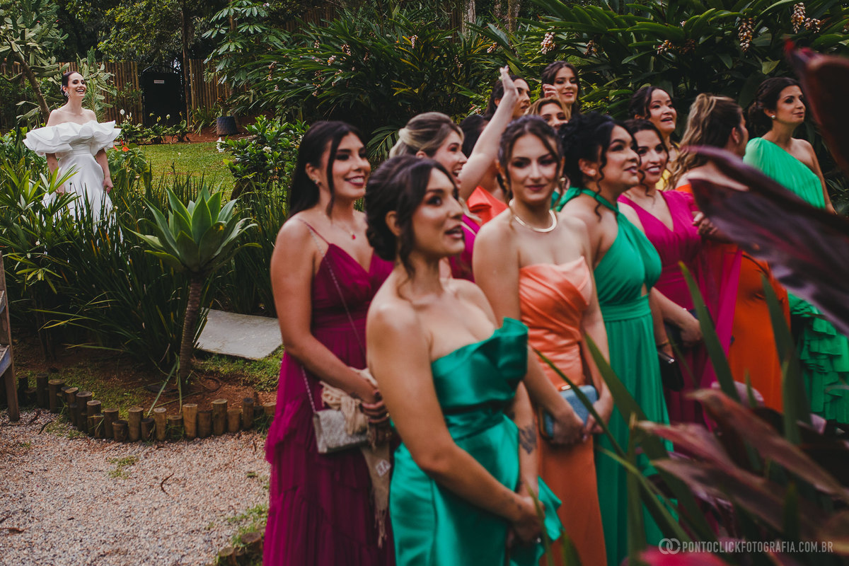Noiva caminha pelo jardim com vestido branco de babados enquanto madrinhas em vestidos vibrantes a acompanham. Foto destaca o contraste de cores e a beleza natural do local