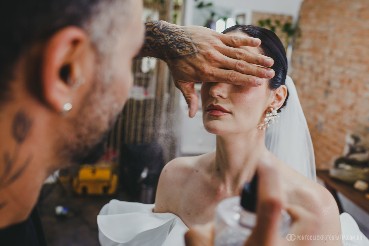 Maquiador aplicando spray fixador no rosto da noiva, dando os toques finais no visual antes da cerimônia. O momento captura a atenção aos detalhes e a preparação cuidadosa para o grande dia