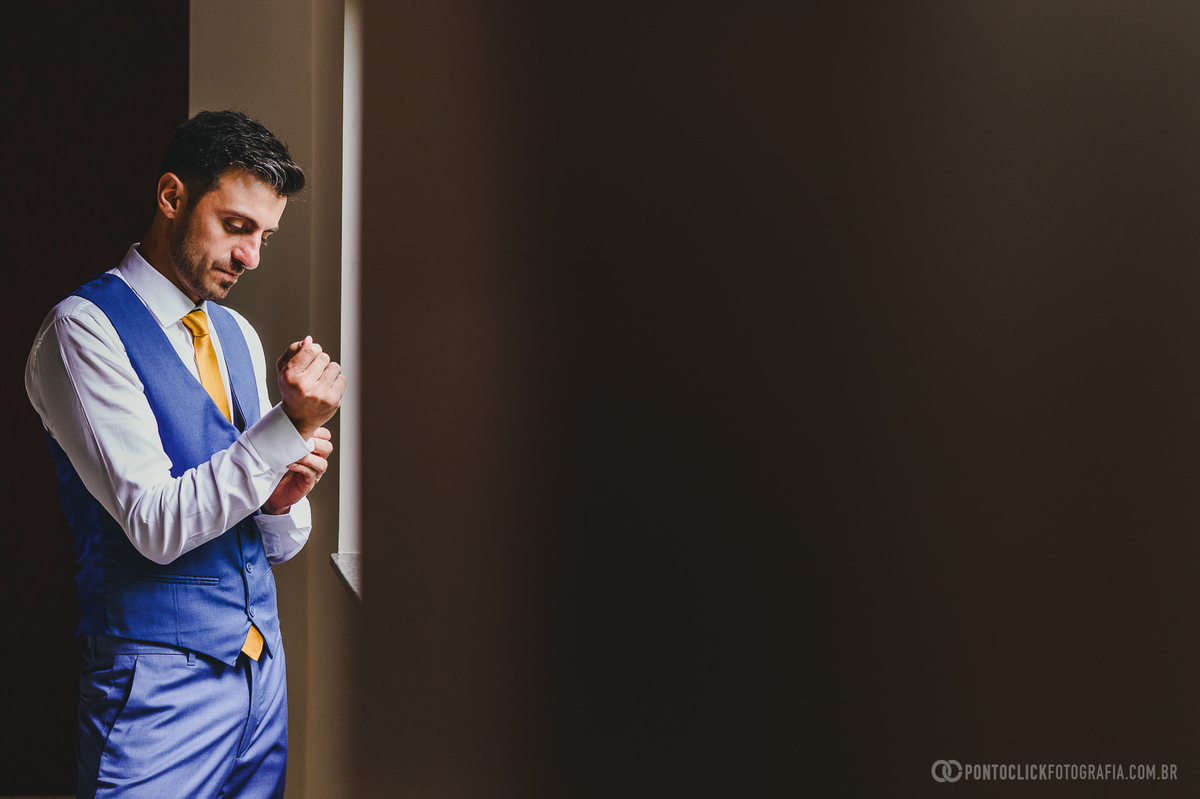 Noivo ajustando os punhos da camisa, com ternos azuis e gravata amarela, antes da cerimônia de casamento. O momento captura a preparação e o foco nos detalhes, com a luz suave que destaca as especificidades e a concentração