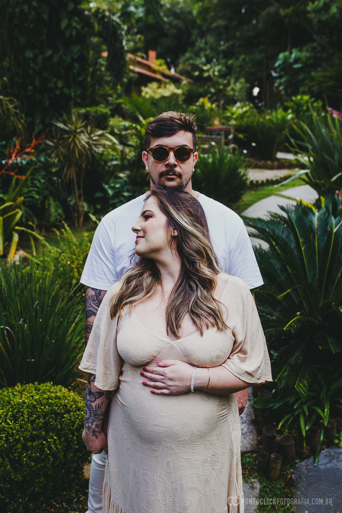 Fernanda e Armando em um retrato sereno, onde ela está protegida com ternura segurando a barriga, enquanto ele a acompanha com uma postura protetora. O cenário verde e exuberante ao fundo reforçam o clima de amor e expectativa