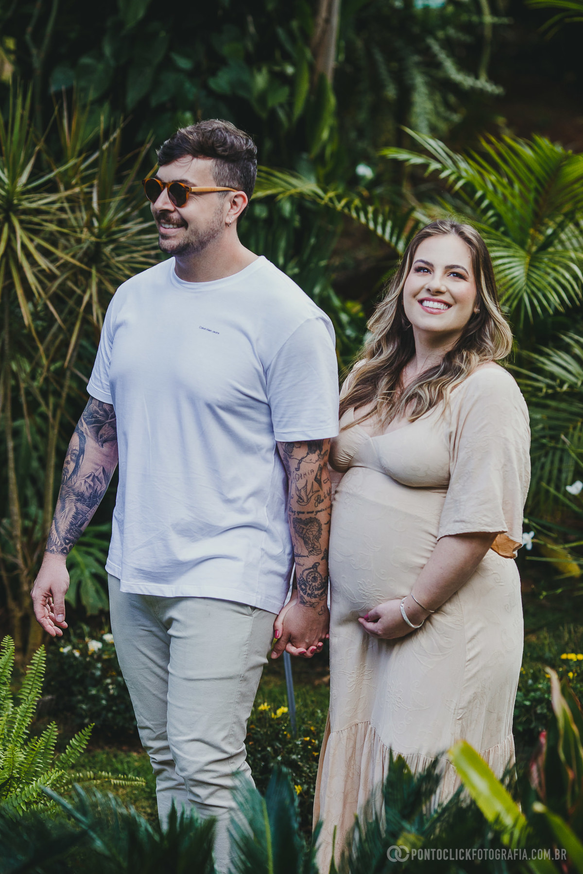 Fernanda e Armando caminham pelo jardim, de mãos dadas, trocando sorrisos cúmplices. O cenário vibrante realça a felicidade do casal.