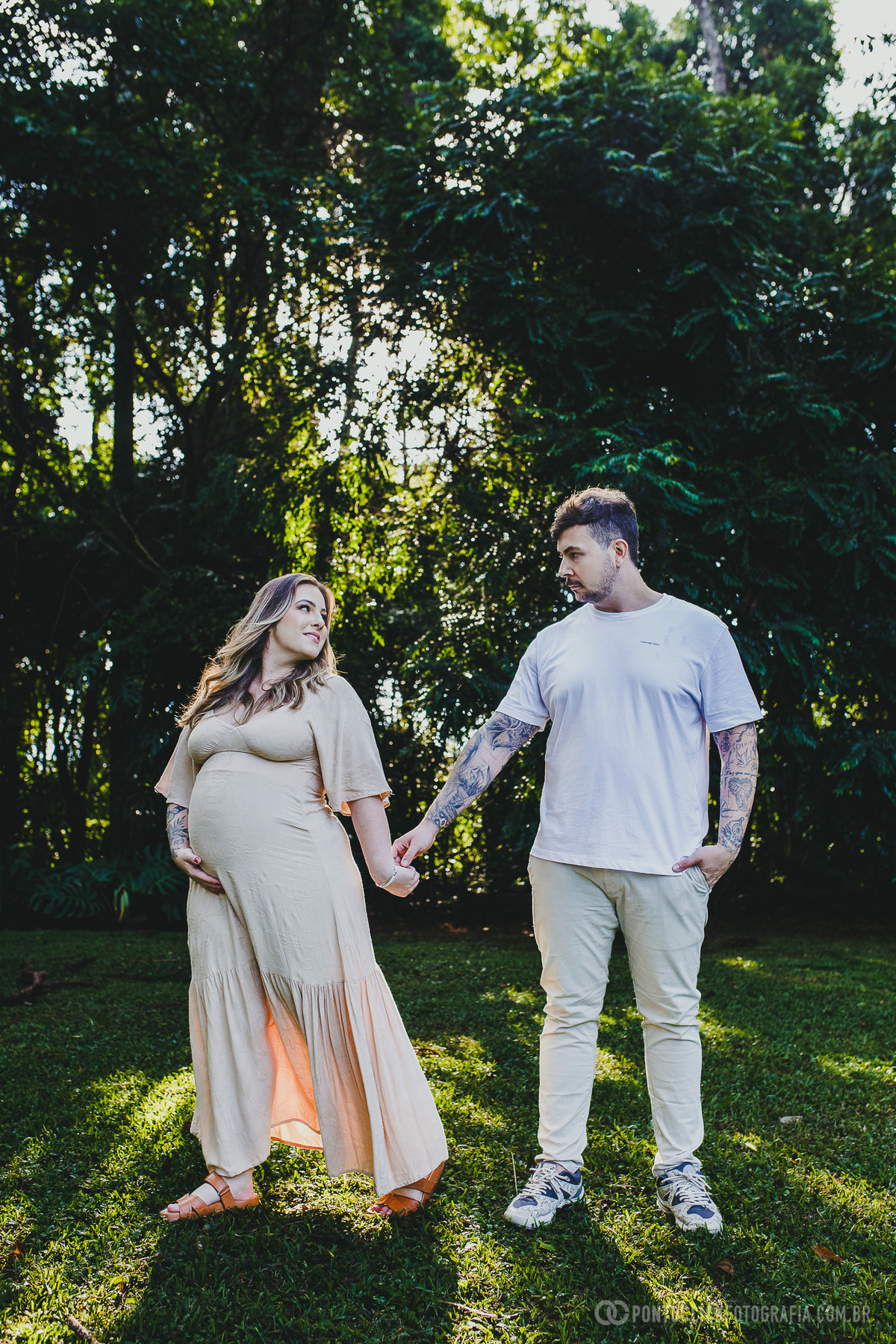 Casal conectado em um ensaio gestante ao ar livre, de mãos dadas em meio a um cenário natural exuberante. A interação entre eles reflete cumplicidade e amor