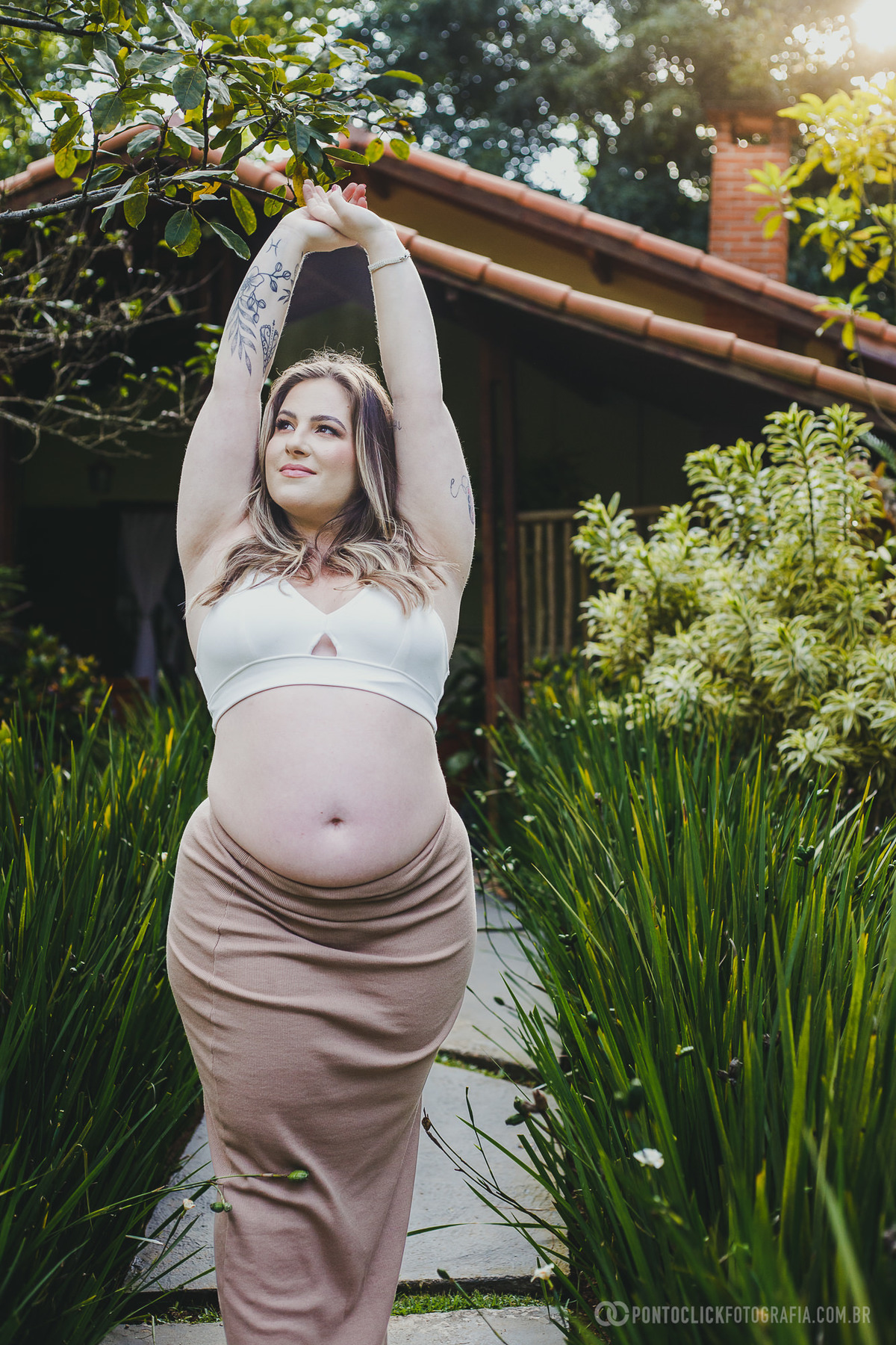 Gestante em pose elegante durante ensaio ao ar livre, com os braços erguidos em meio ao verde vibrante da vegetação. O visual destaca sua confiança e conexão com a maternidade.