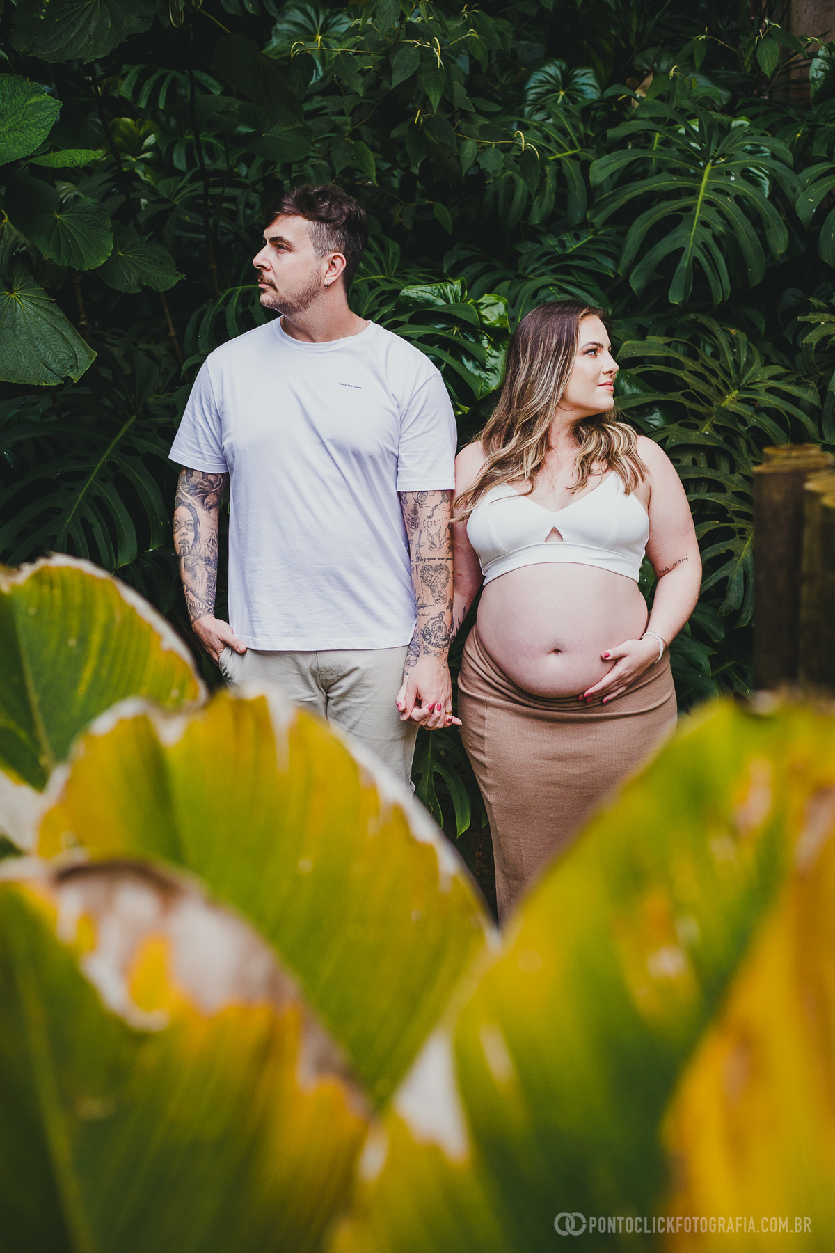 Casal em pose descontraída durante ensaio de gestante, rodeado por folhas tropicais que realçam a composição visual e reforçam a essência natural do momento.