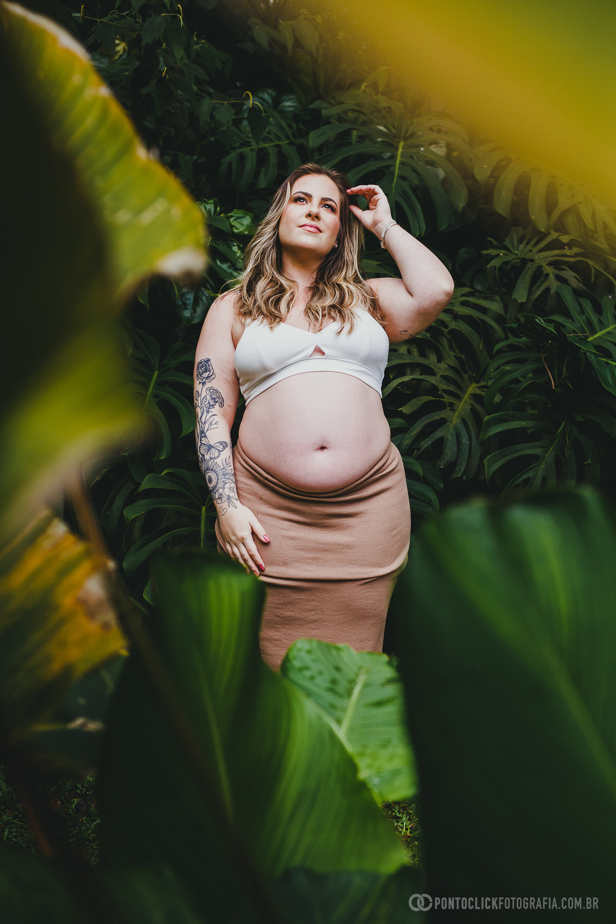 Retrato de gestante em meio a folhas tropicais, capturando um olhar contemplativo e sereno. A fotografia destaca a beleza da maternidade em harmonia com a natureza.