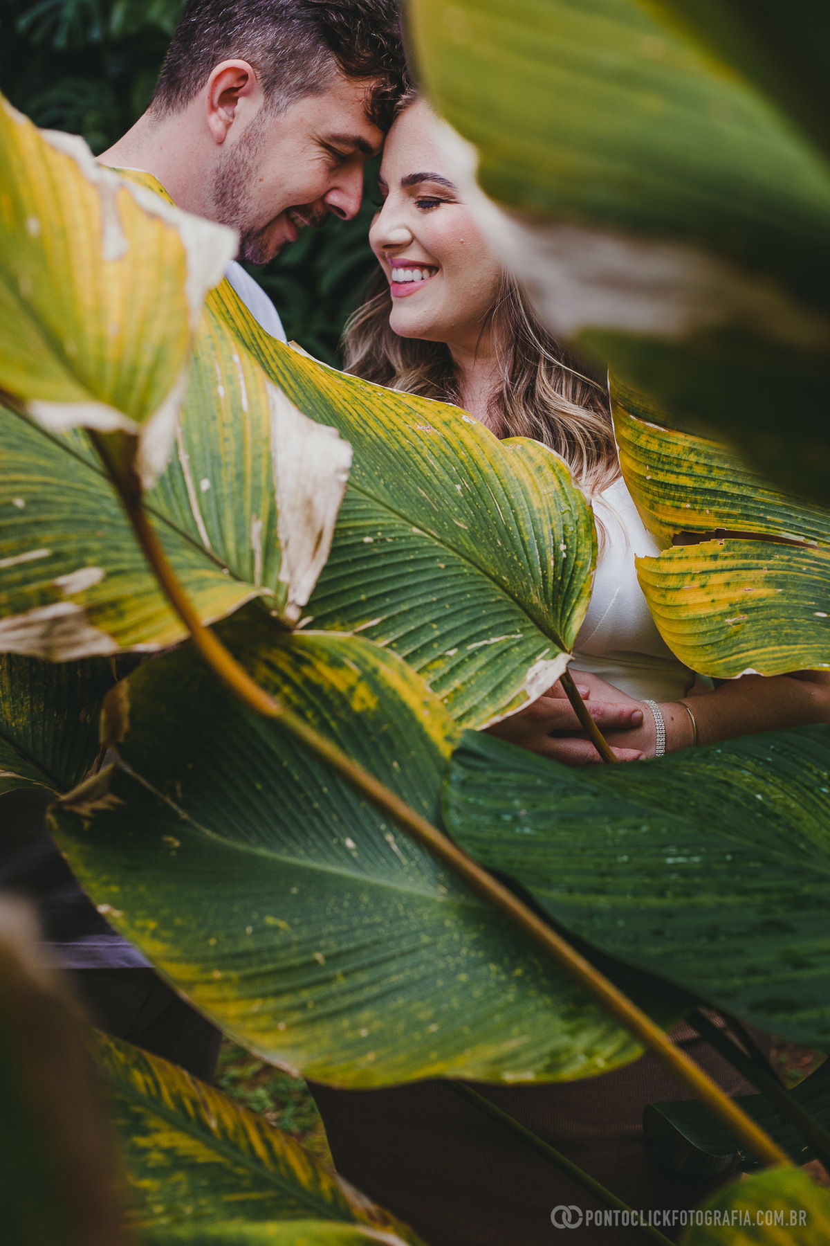 Close de casal em ensaio gestante, compartilhando um momento íntimo e carinhoso em meio às folhas tropicais. A composição enfatiza a conexão e o amor