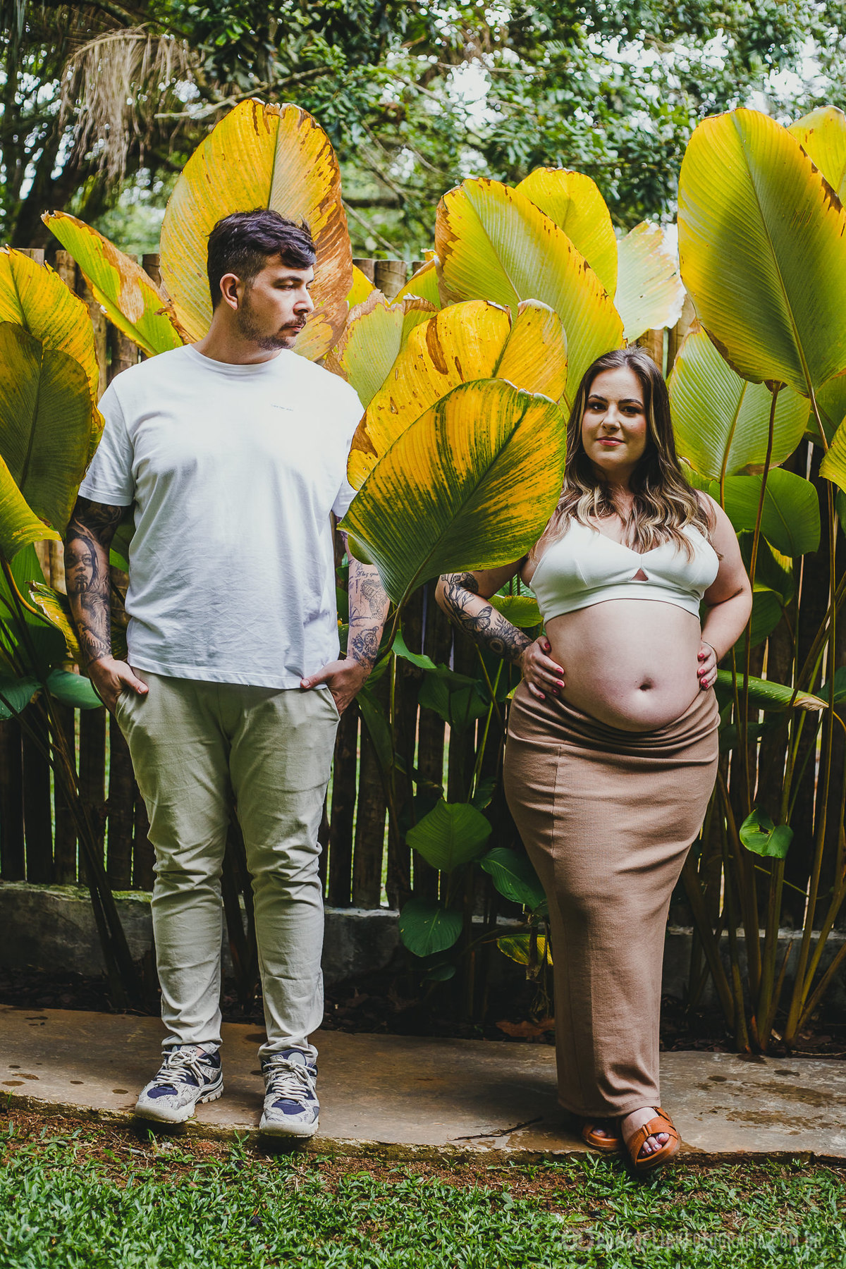 Casal lado a lado em ensaio gestante, posicionados em meio a uma folhagem tropical vibrante. Uma fotografia captura a harmonia entre eles e o cenário natural.
