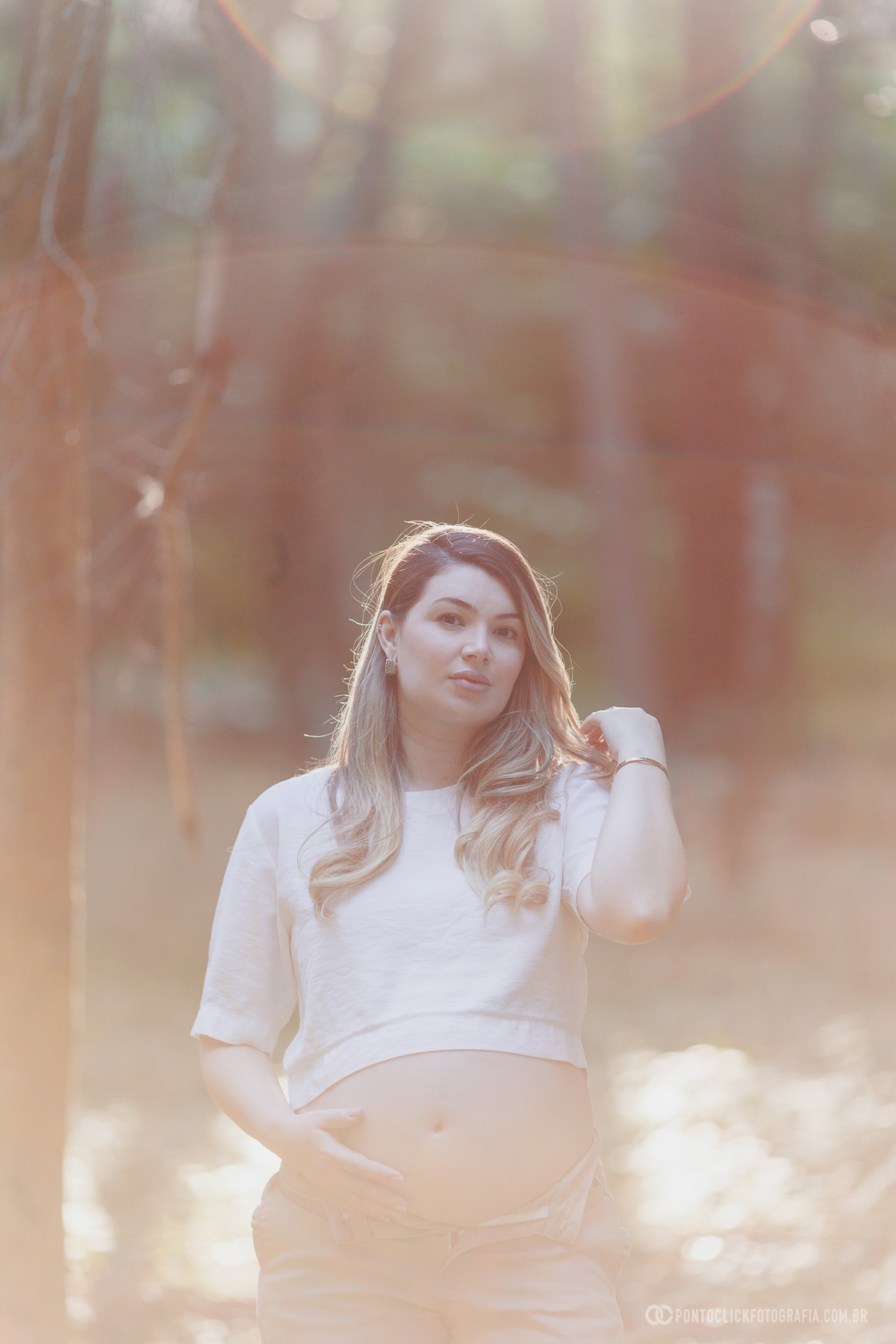 ensaio gestante com luz contra o sol criando flare suave destacando a gestante com expressão serena e conexão com a maternidade em ambiente natural em São Roque