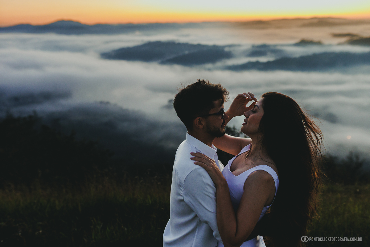 Casal em ensaio pré wedding no Morro do Capuava em Pirapora do Bom Jesus caminhando próximo à bandeira com céu colorido ao amanhecer e vegetação ao redor compondo cenário natural