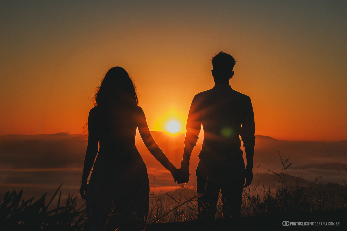 Casal em ensaio pré wedding no Morro do Capuava em Pirapora do Bom Jesus caminhando juntos com mar de nuvens ao fundo durante nascer do sol, criando composição ampla com luz suave e atmosfera romântica