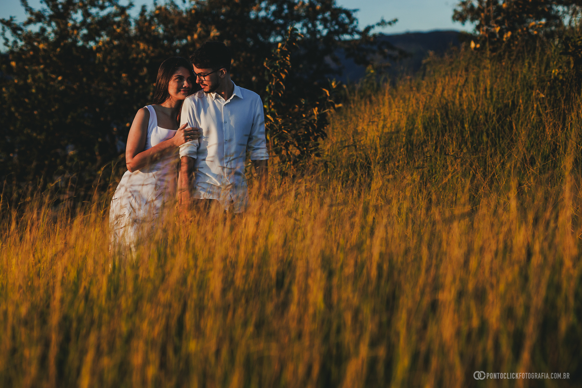 Casal em ensaio pré wedding no Morro do Capuava em Pirapora do Bom Jesus caminhando de mãos dadas com bandeira ao lado e céu em tons suaves ao amanhecer, criando cenário natural com leveza e movimento