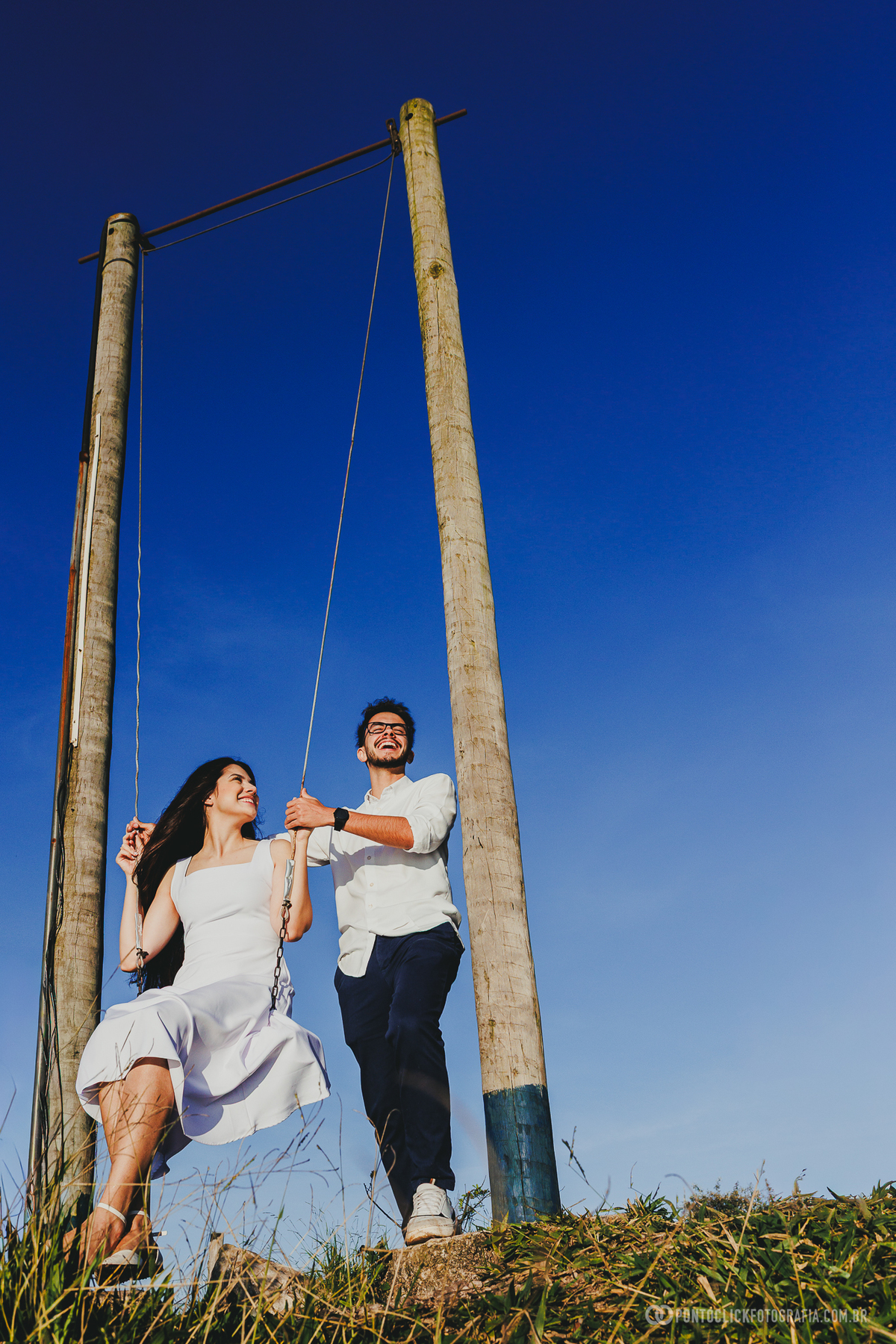Casal em ensaio pré wedding no Morro do Capuava em Pirapora do Bom Jesus em balanço de madeira com céu azul limpo ao fundo criando cena leve, divertida e natural ao ar livre