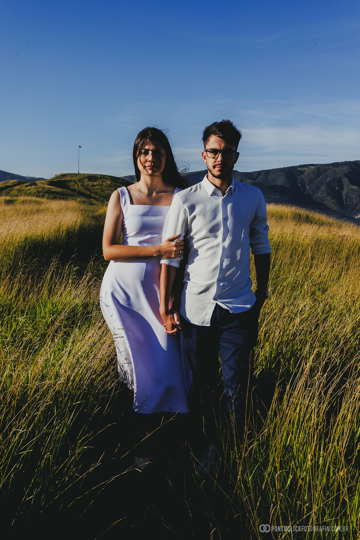 Casal em ensaio pré wedding no Morro do Capuava em Pirapora do Bom Jesus caminhando entre vegetação alta com luz lateral e montanhas ao fundo criando composição natural
