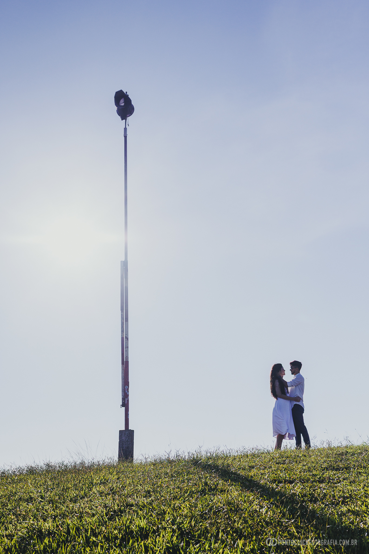 Casal em ensaio pré wedding no Morro do Capuava em Pirapora do Bom Jesus caminhando em linha com bandeira ao centro e céu limpo criando composição vertical equilibrada
