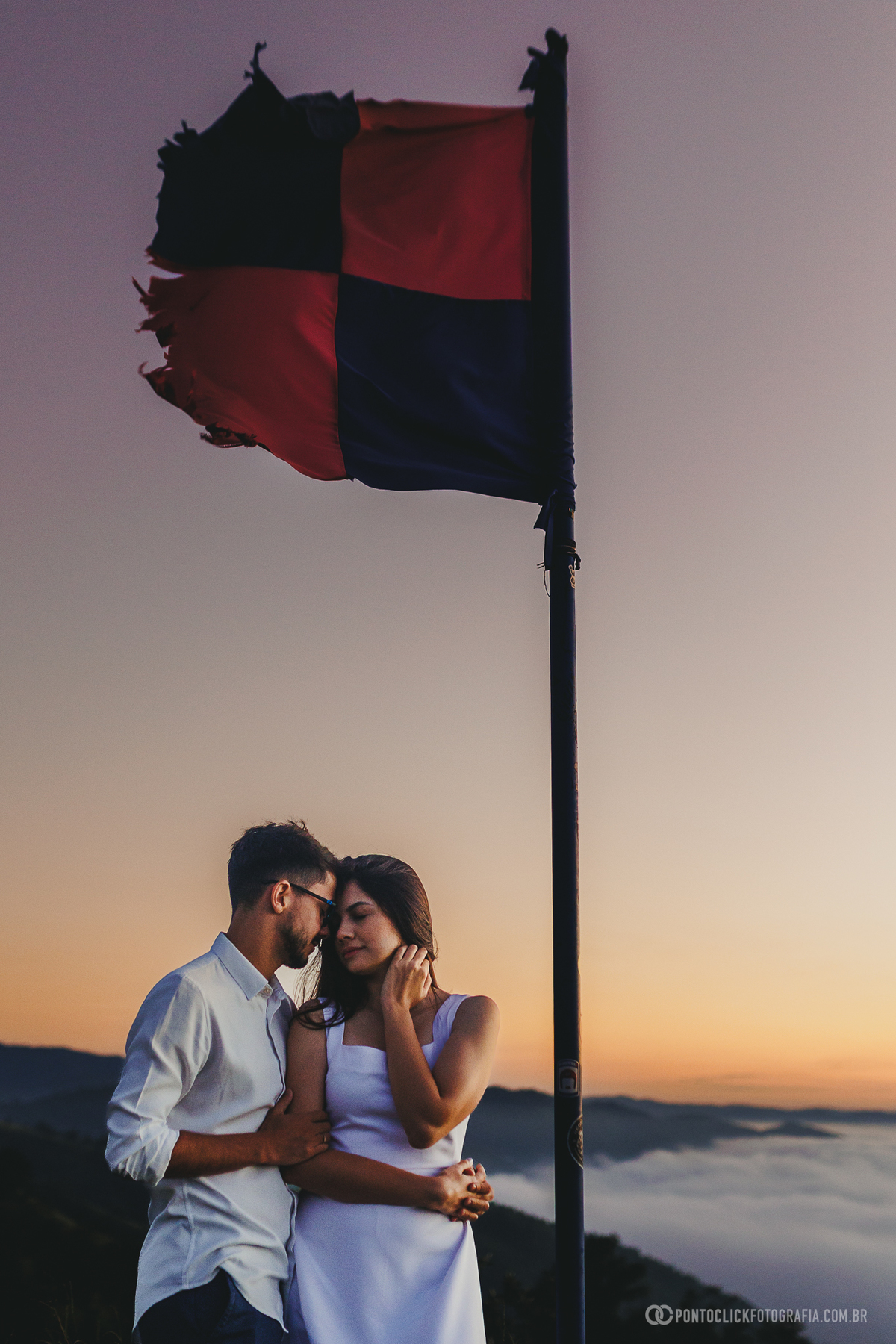 Bandeira em destaque no Morro do Capuava em Pirapora do Bom Jesus durante nascer do sol com céu limpo e luz suave criando contraste com o cenário natural e mar de nuvens ao fundo
