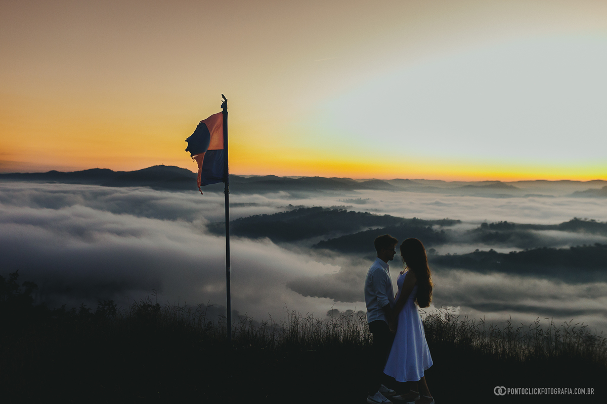 Casal em ensaio pré wedding no Morro do Capuava em Pirapora do Bom Jesus com bandeira ao lado e mar de nuvens ao fundo durante nascer do sol, criando cenário romântico com luz natural