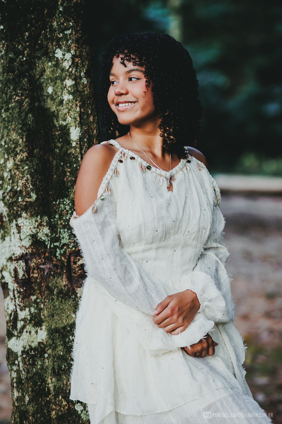 Ensaio feminino de 15 anos em São Roque no Brasital com debutante apoiada em árvore, vestindo branco, em fotografia com luz suave, sorriso espontâneo e fundo verde desfocado, transmitindo naturalidade, charme e leveza no retrato