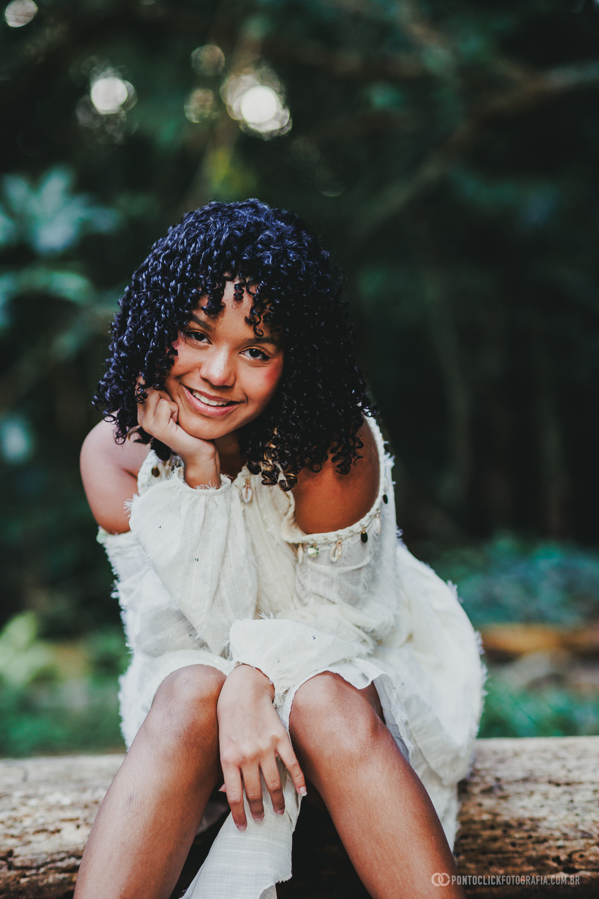 Retrato de ensaio de 15 anos no Brasital em São Roque com debutante sorrindo e apoiando o rosto na mão, usando vestido branco, em fotografia delicada com fundo verde desfocado valorizando doçura, juventude e expressão natural