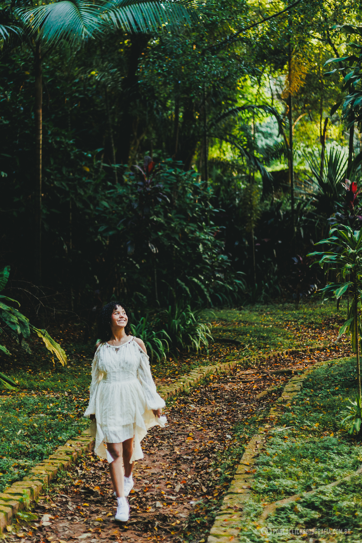 Fotografia de debutante em ensaio de 15 anos no Brasital em São Roque com jovem caminhando por trilha cercada por natureza, usando vestido branco, em composição ampla que destaca cenário natural, leveza e beleza do local