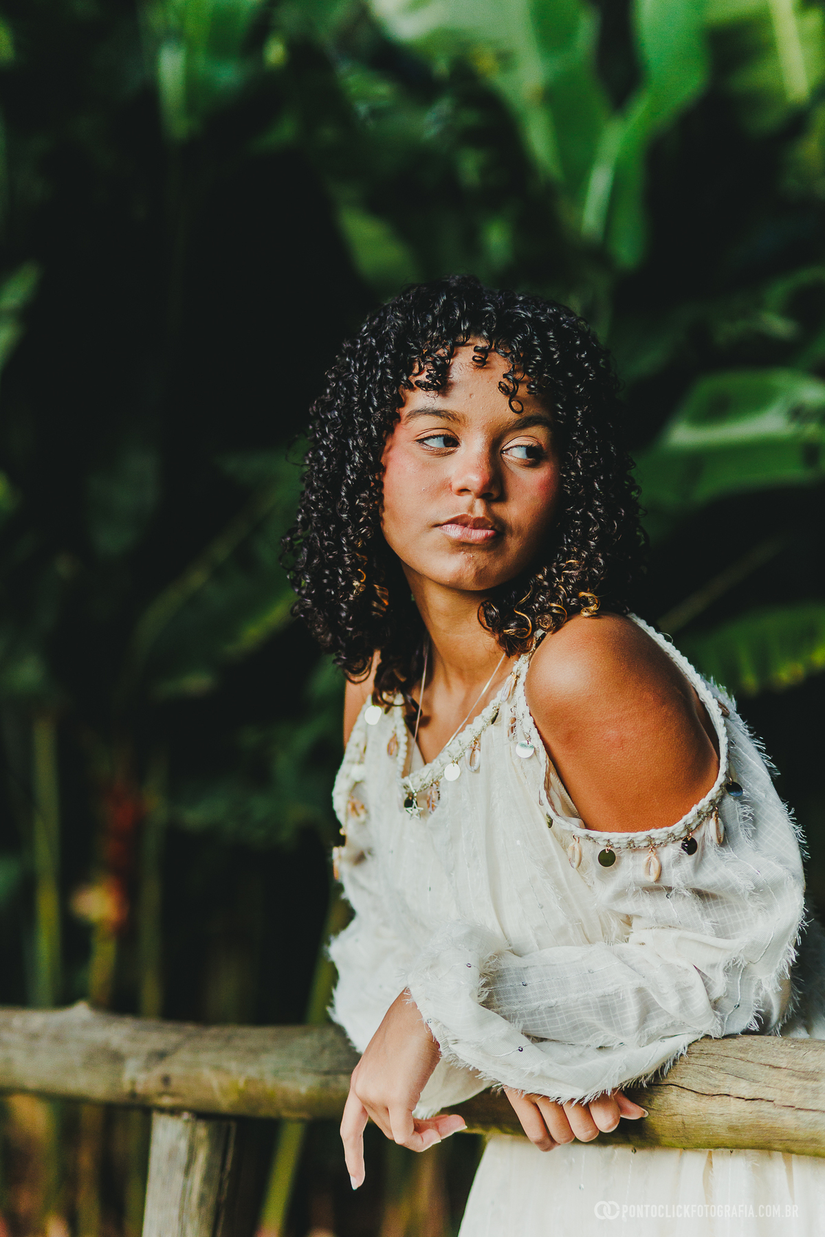 Retrato de ensaio de 15 anos em São Roque no Brasital com debutante apoiada em cerca de madeira, vestindo branco, em fotografia com fundo verde intenso e luz natural suave destacando elegância e expressão marcante