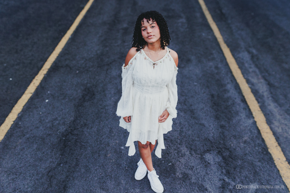 Ensaio feminino de 15 anos em São Roque com debutante em pé sobre asfalto, usando vestido branco, em fotografia com composição simétrica e linhas no chão, destacando estilo, atitude e beleza em cenário urbano contemporâneo