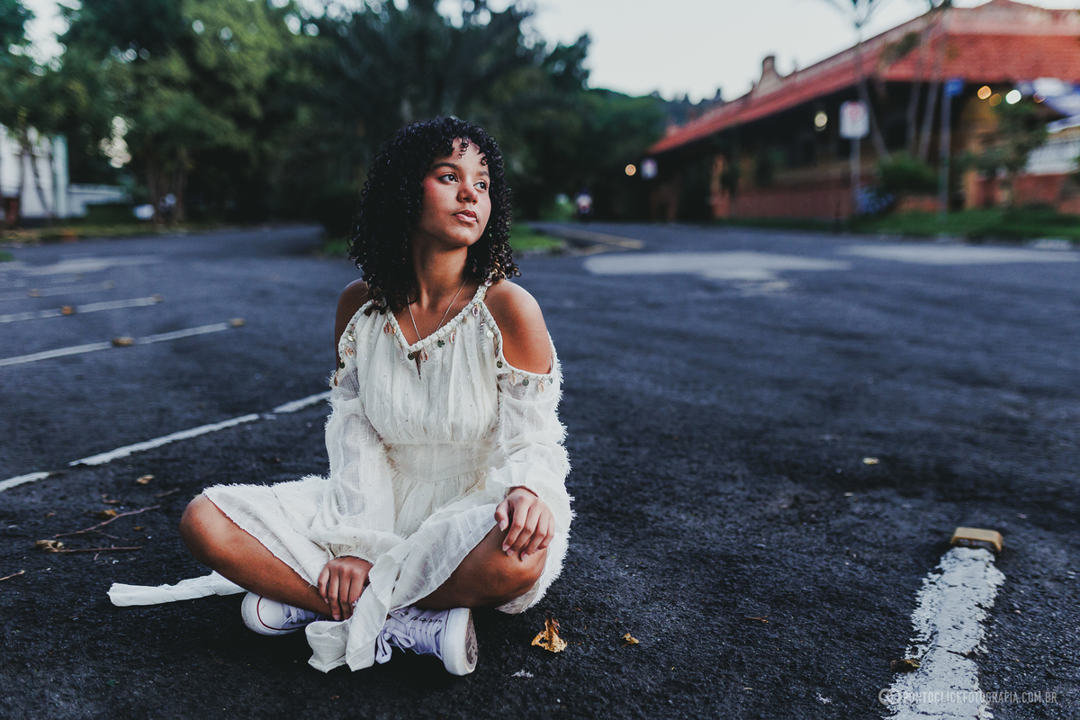 Retrato de debutante em ensaio de 15 anos em São Roque com jovem sentada no asfalto, usando vestido branco, em composição urbana com fundo desfocado, transmitindo personalidade, delicadeza e contraste com o ambiente