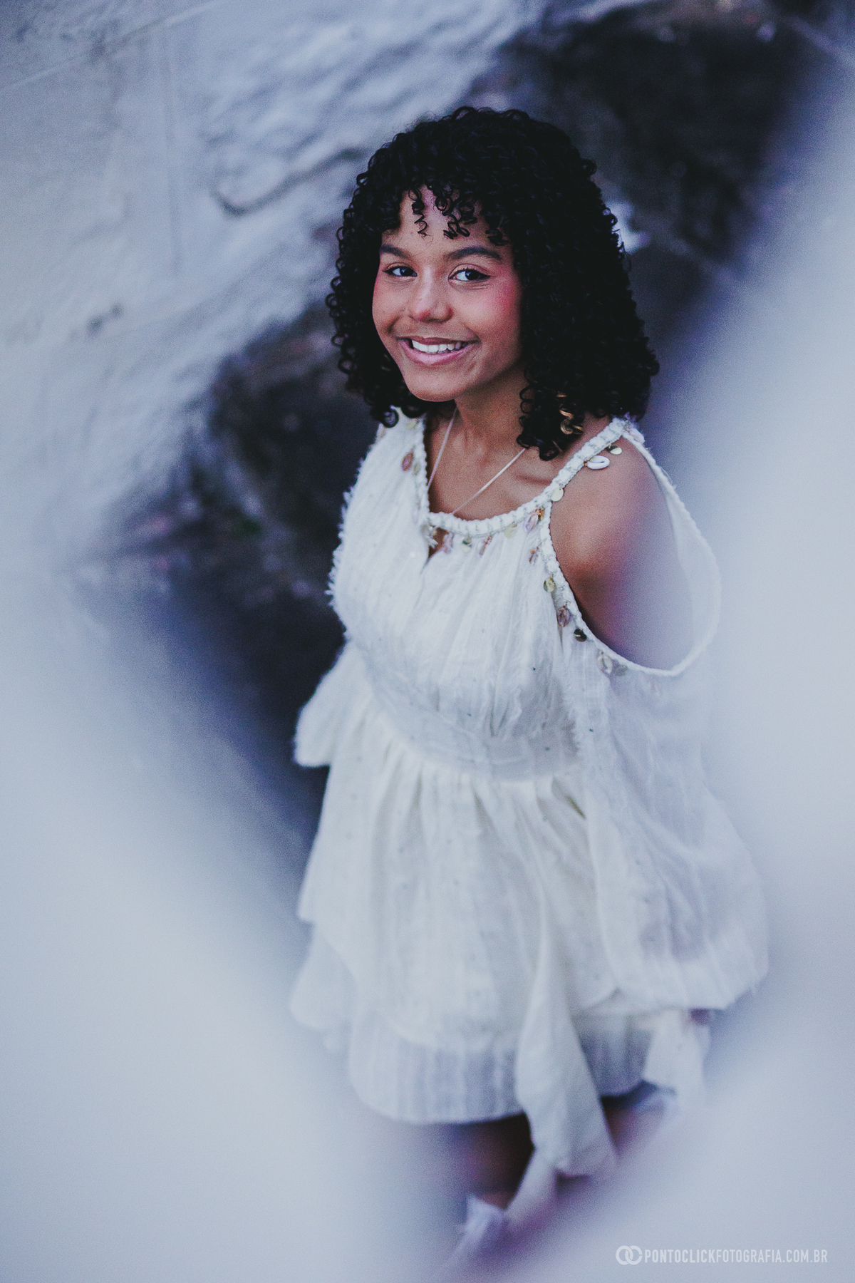 Fotografia artística de ensaio de 15 anos em São Roque no Brasital com debutante em vestido branco, sorriso natural e desfoque em primeiro plano, criando composição delicada que valoriza expressão e sensibilidade