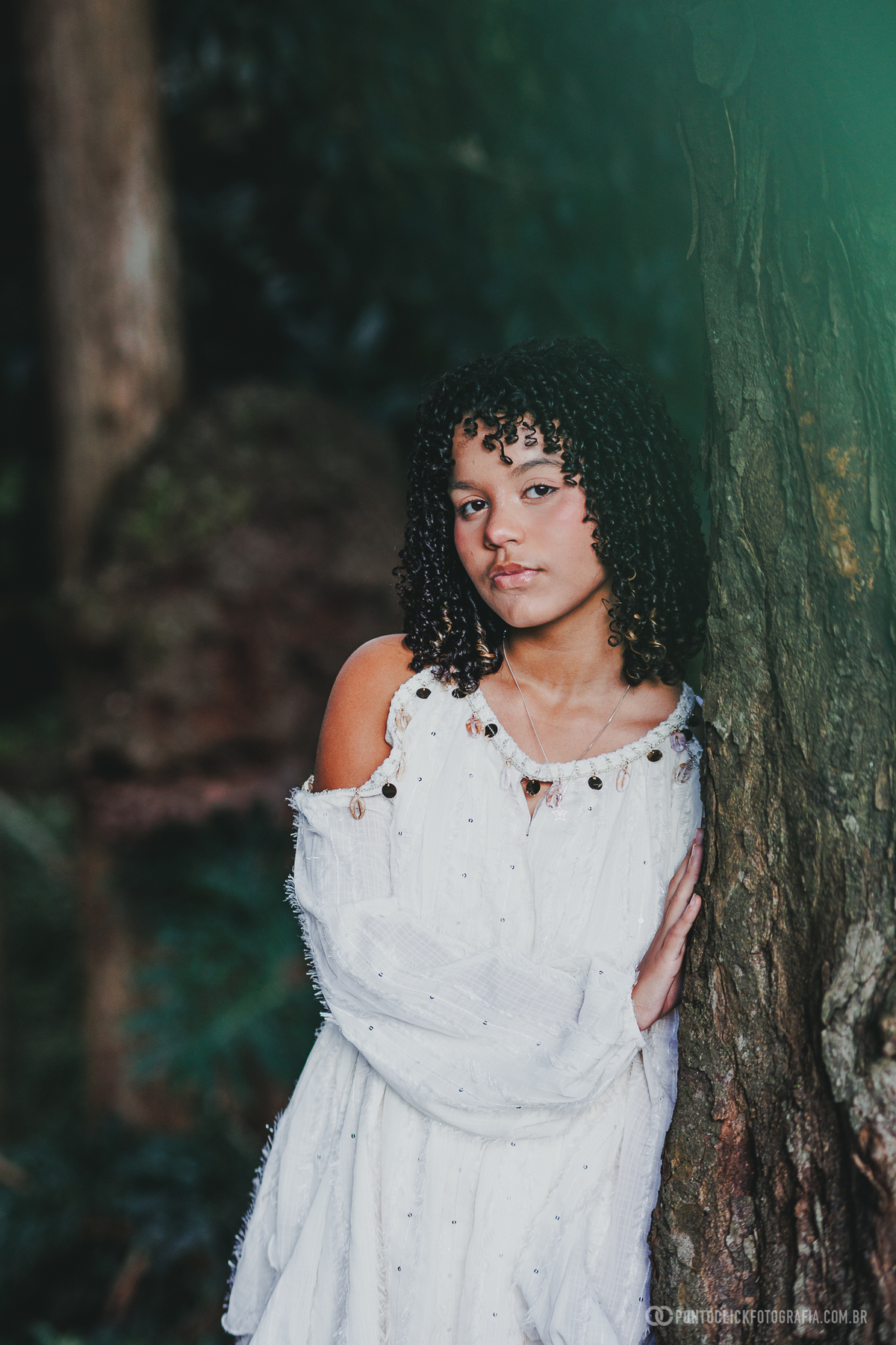 Retrato de ensaio de 15 anos no Brasital em São Roque com debutante apoiada em árvore, vestido branco fluido e olhar marcante, fotografia com fundo verde desfocado e luz suave, destacando elegância, delicadeza e conexão com a natureza