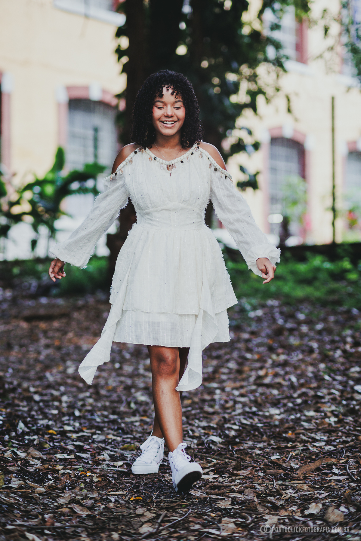 Ensaio de debutante de 15 anos em São Roque no Brasital com jovem caminhando sorrindo, vestido branco e folhas no chão, fotografia espontânea com fundo desfocado transmitindo leveza, alegria e naturalidade em momento ao ar livre