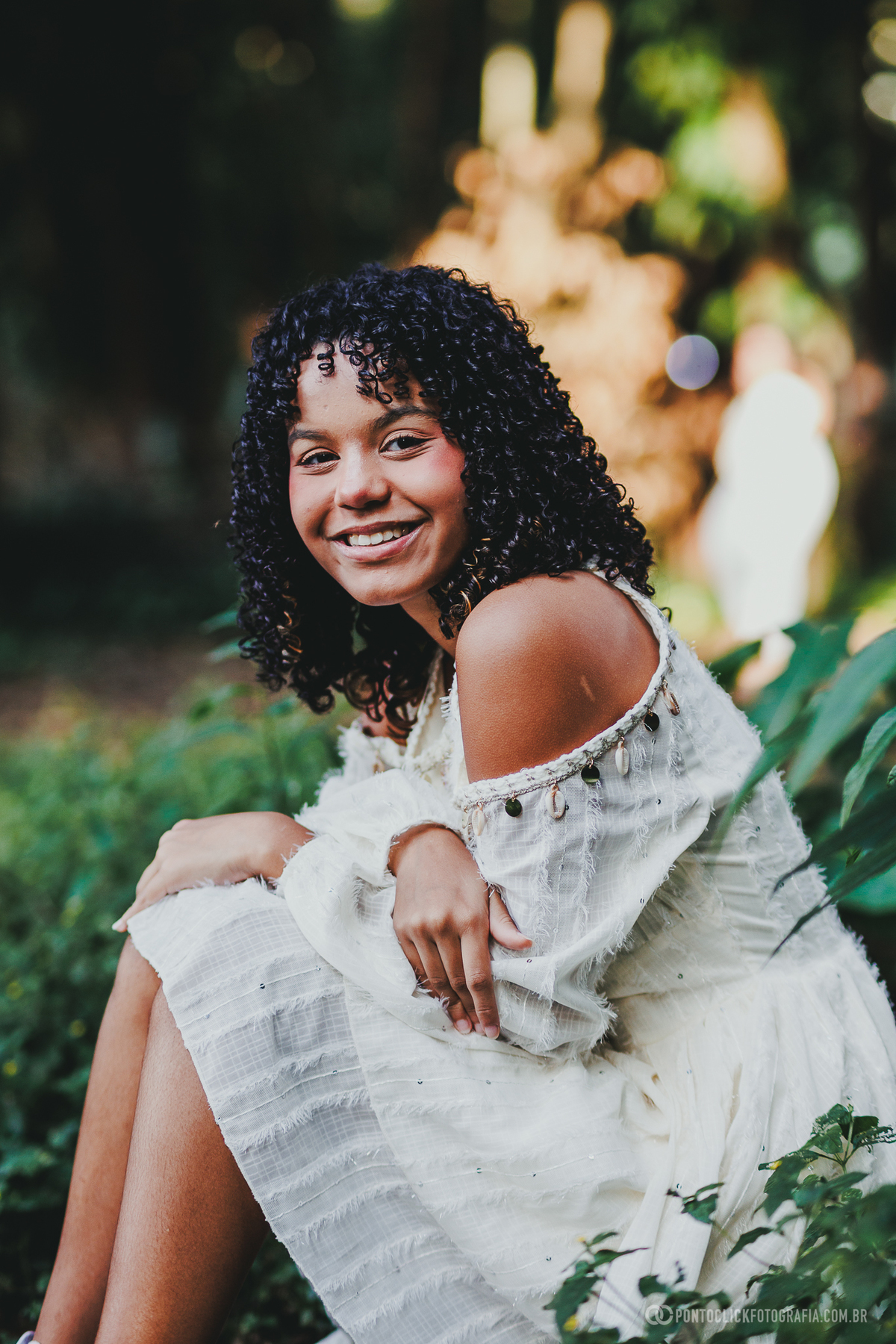 Ensaio feminino de 15 anos em São Roque no Brasital com debutante sorrindo sentada em meio à natureza, vestido branco e fotografia espontânea com fundo desfocado e luz suave, transmitindo alegria, autenticidade e leveza no cenário
