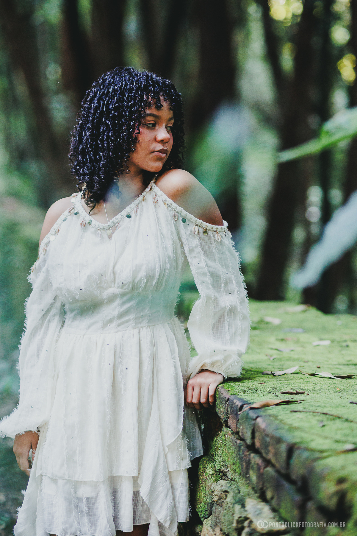 Retrato de debutante em ensaio de 15 anos no Brasital em São Roque com jovem ao lado de estrutura de pedra com musgo, usando vestido branco, em composição delicada com fundo verde desfocado e atmosfera natural elegante e suave