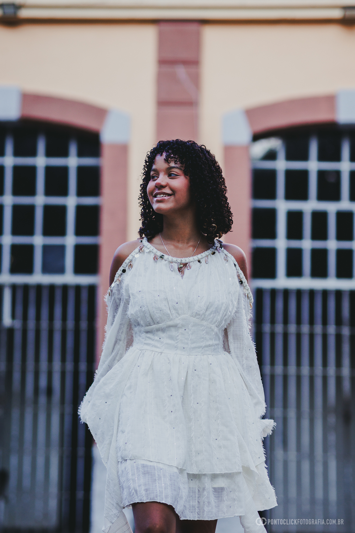 Retrato de debutante em ensaio de 15 anos no Brasital em São Roque com vestido branco e sorriso leve diante de arquitetura desfocada, em composição elegante que destaca juventude, delicadeza e beleza natural em ambiente externo