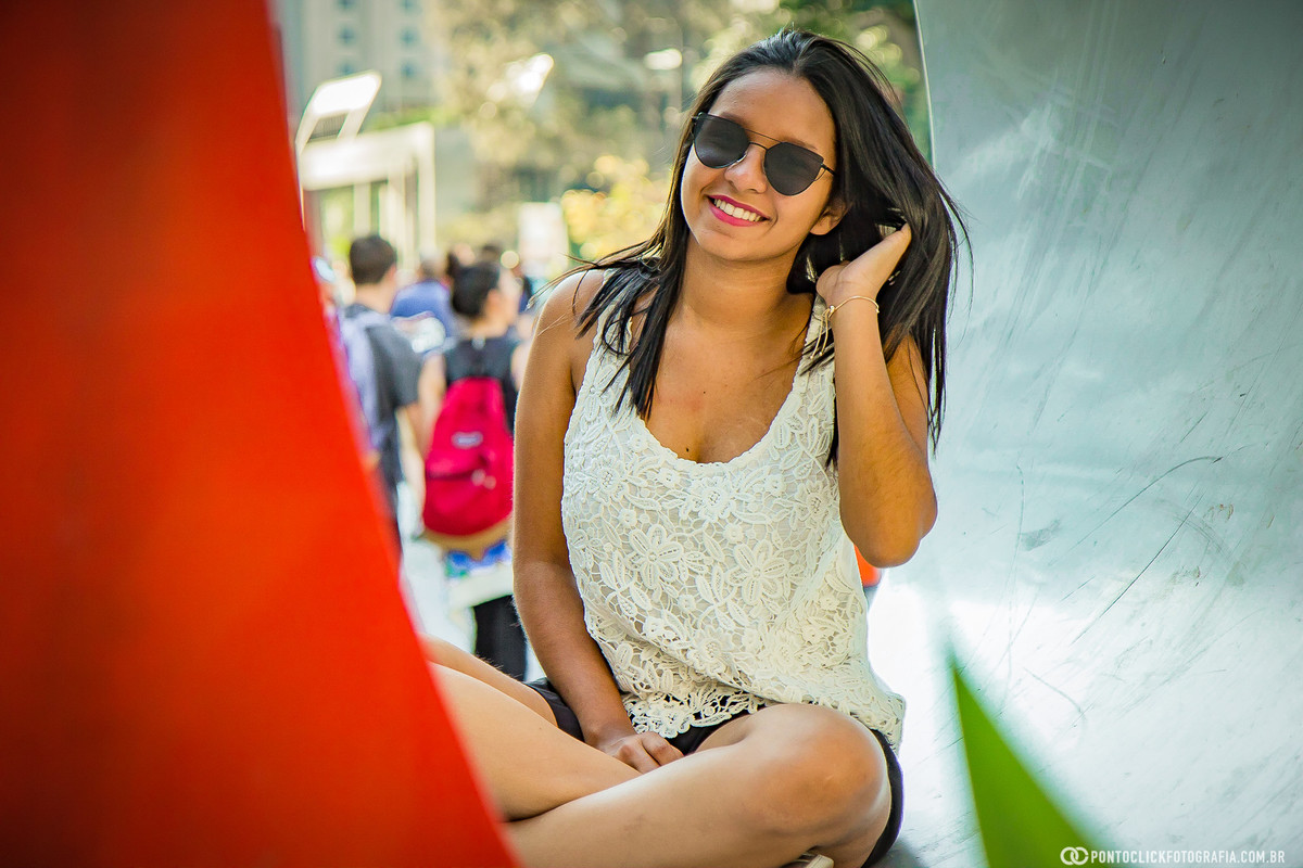 sorrindo e com a mão no cabelo e sentada na escultura da avenida paulista