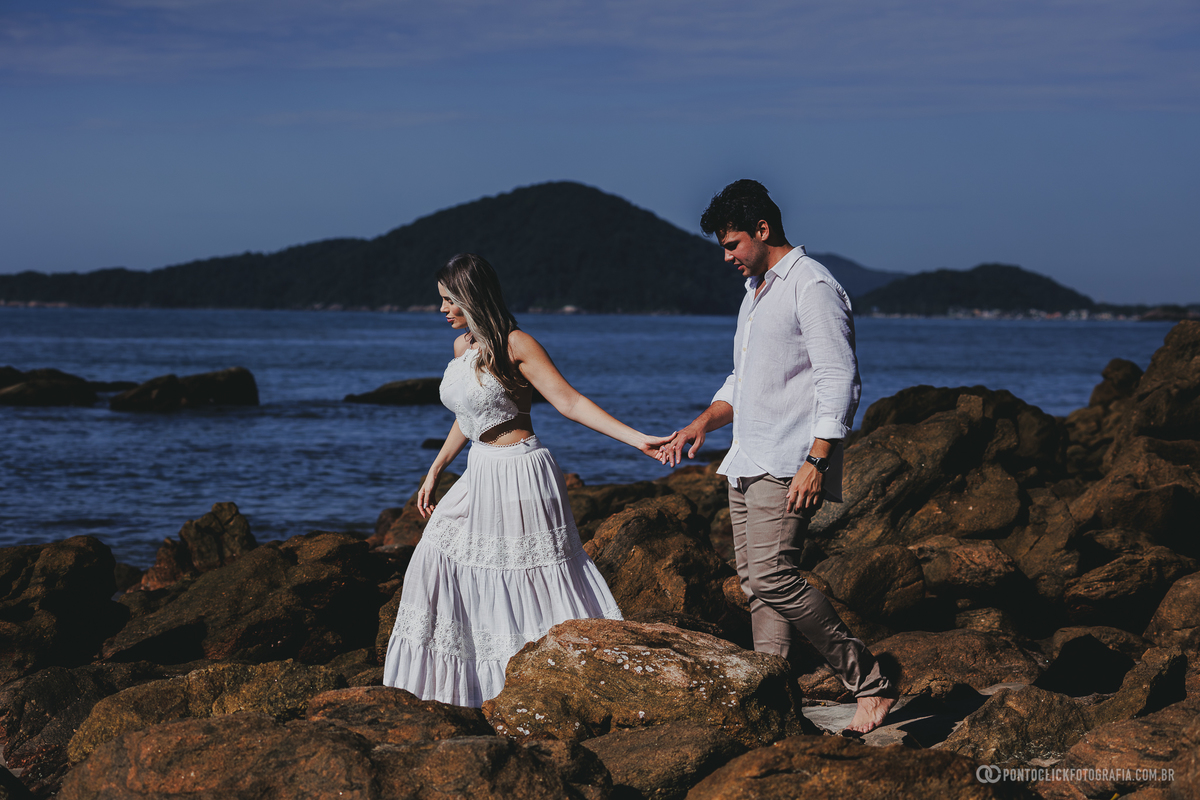 Fotografia de casal realizada no Guarujá com Guilherme e Katioska caminhando entre as pedras da praia, destacando conexão, espontaneidade e cenário natural do litoral paulista durante o ensaio pré casamento