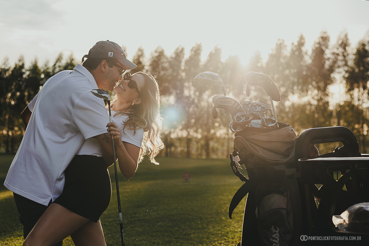 Casal troca um momento de carinho ao lado dos equipamentos de golfe durante ensaio fotográfico lifestyle. A composição reúne luz natural, espontaneidade e um cenário elegante para registrar a conexão entre os dois