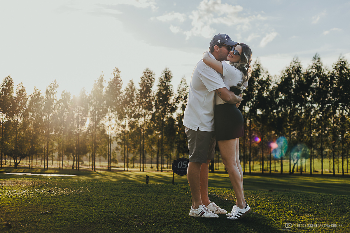 Casal se abraça durante ensaio fotográfico em campo de golfe, aproveitando a luz do fim da tarde para criar imagens naturais e cheias de emoção. O cenário reforça a proposta elegante e descontraída do ensaio de casal