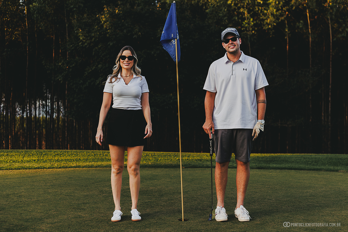 Casal posa ao lado da bandeira do green durante ensaio fotográfico em campo de golfe. A fotografia valoriza elegância, sintonia e um cenário sofisticado ao ar livre, criando imagens naturais para ensaio lifestyle de casal