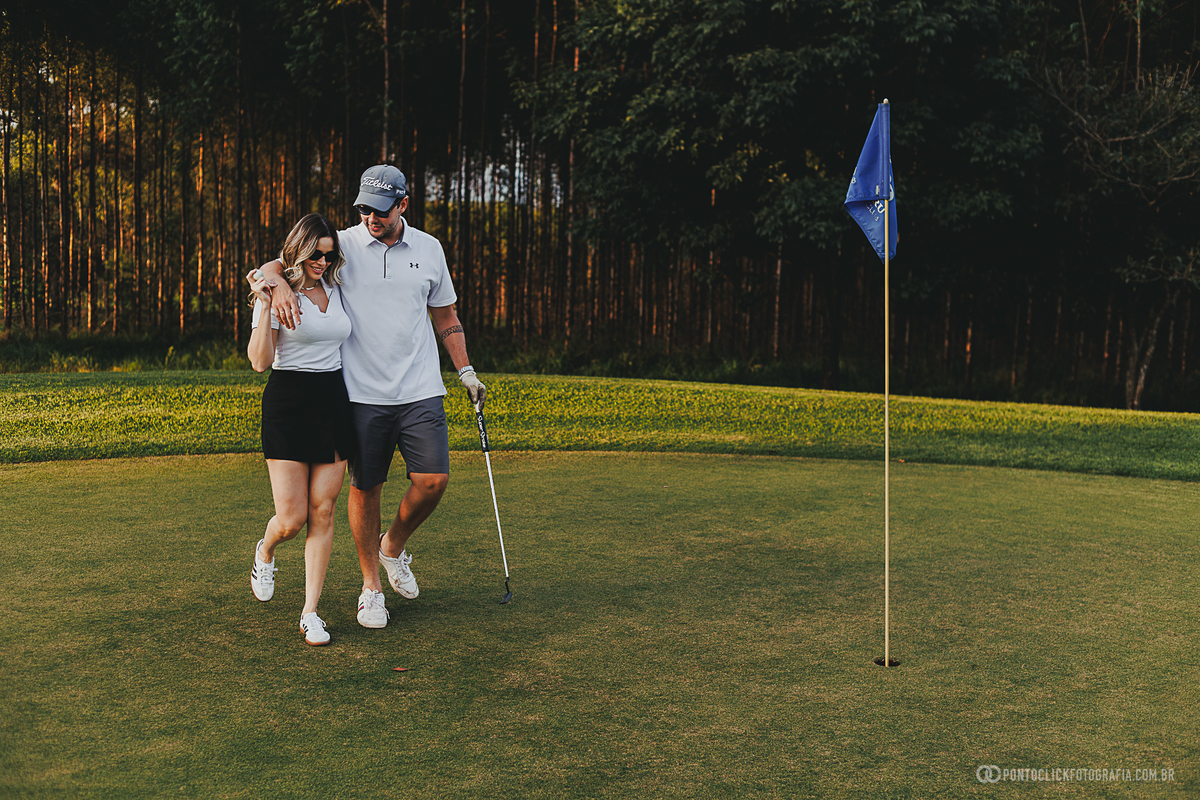Casal caminha abraçado pelo green durante ensaio fotográfico em campo de golfe. A imagem transmite leveza, conexão e espontaneidade, destacando a beleza do cenário natural para um ensaio de casal elegante e atemporal