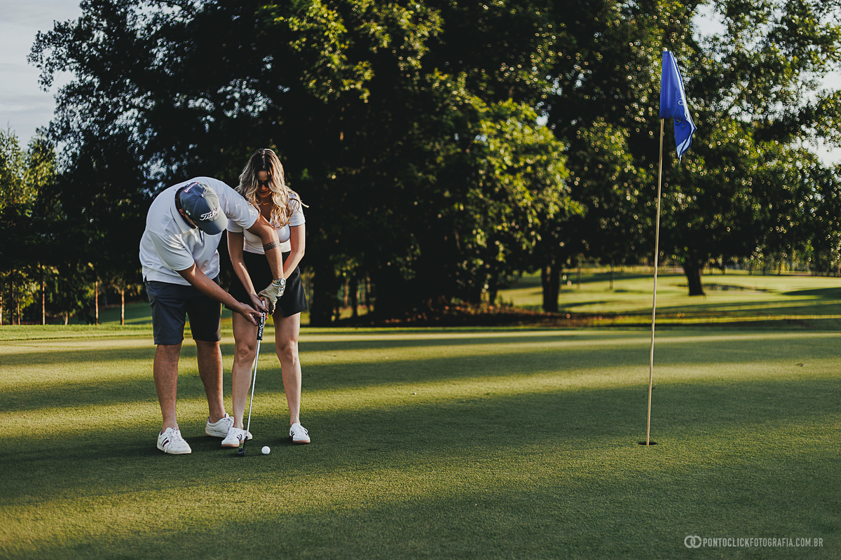 Casal pratica tacadas no green durante ensaio fotográfico em campo de golfe, compartilhando momentos de aprendizado e diversão. A fotografia destaca interação, naturalidade e o cenário perfeito para ensaio lifestyle de casal