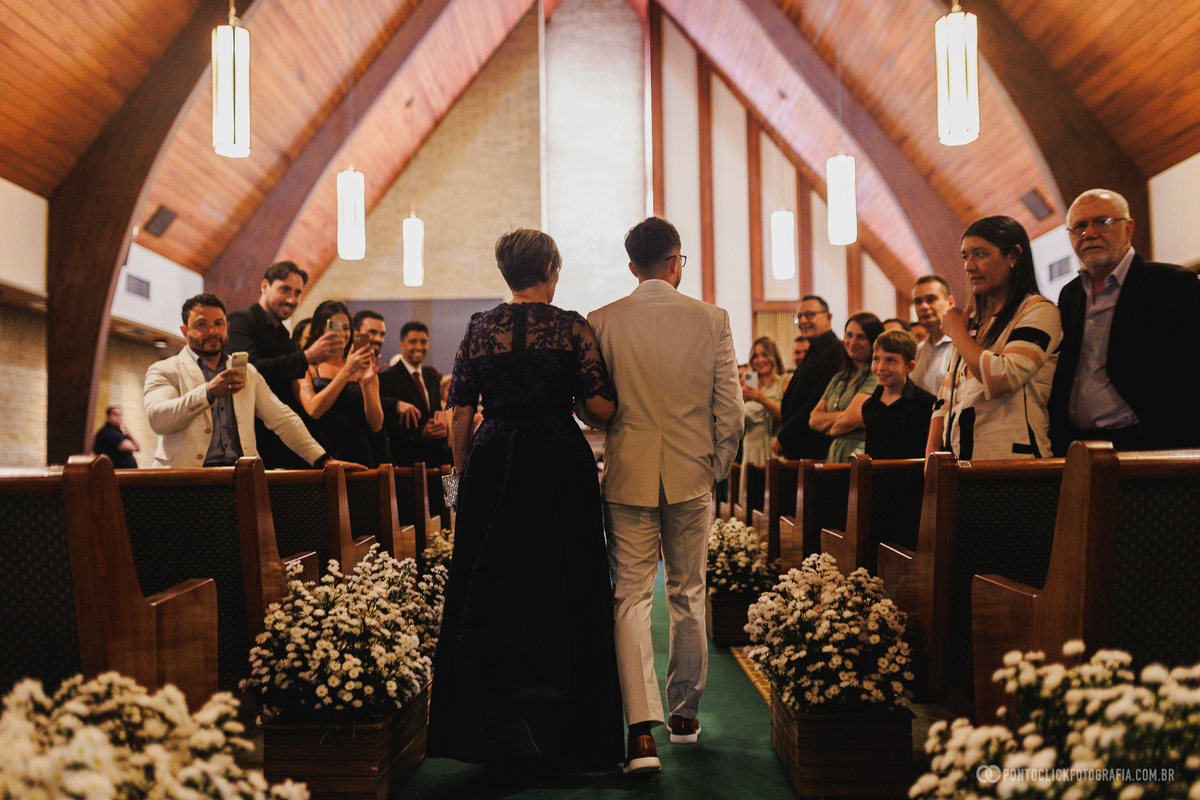 O noivo entra na igreja acompanhado de sua mãe, iniciando a cerimônia de casamento diante de familiares e amigos. Fotógrafo de casamento Pontoclick Fotografia.