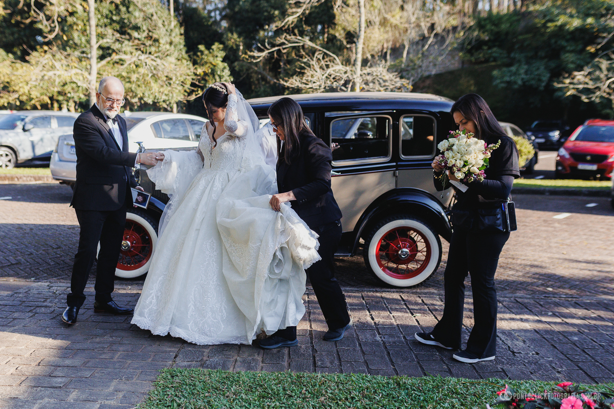 A noiva desce do carro antigo com o auxílio da equipe e do pai, vivendo os instantes finais antes da emocionante entrada na cerimônia de casamento. Fotógrafo de casamento Pontoclick Fotografia.