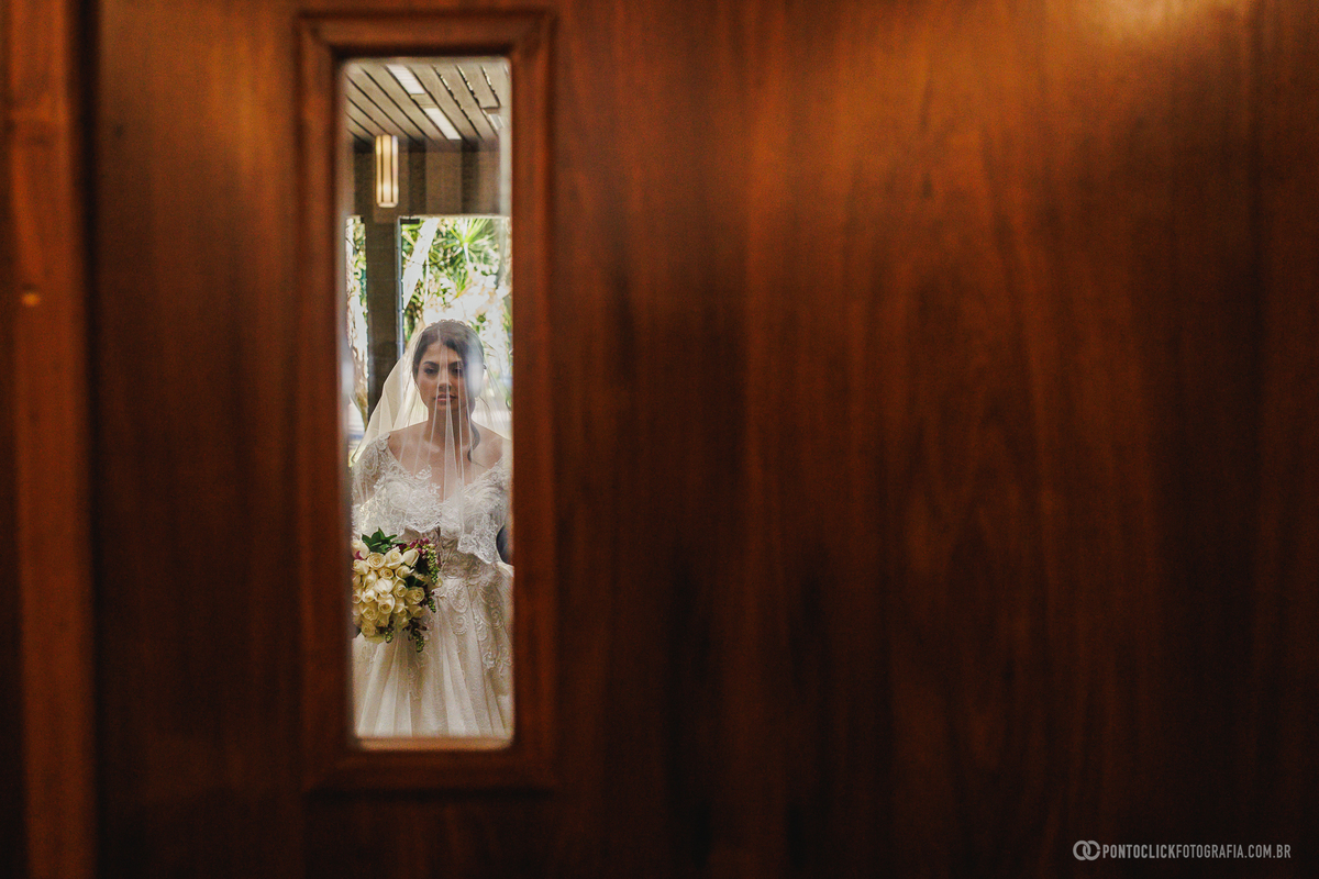 A noiva aguarda o momento da entrada para a cerimônia em um enquadramento criativo, revelando toda a expectativa dos instantes que antecedem o casamento. Fotógrafo de casamento Pontoclick Fotografia