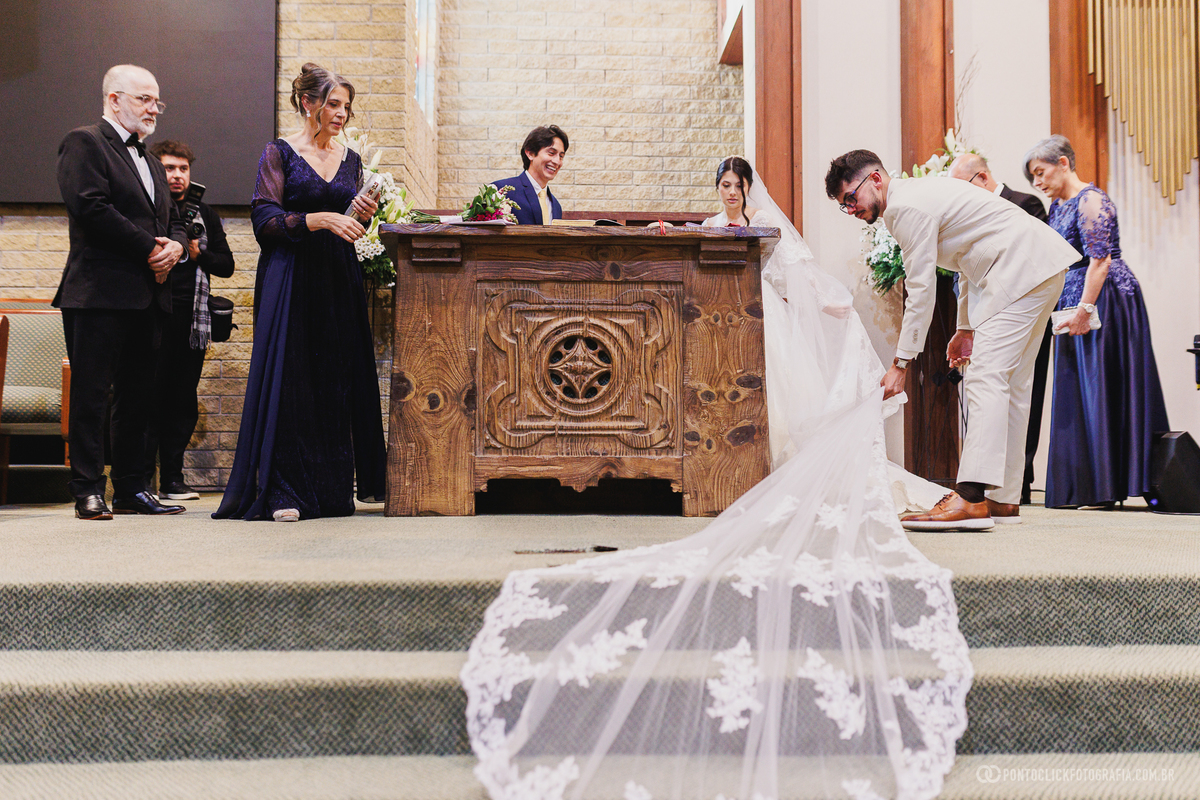 O noivo ajuda a organizar o vestido da noiva diante do altar durante a cerimônia, em um gesto espontâneo de cuidado e carinho. Fotógrafo de casamento Pontoclick Fotografia.