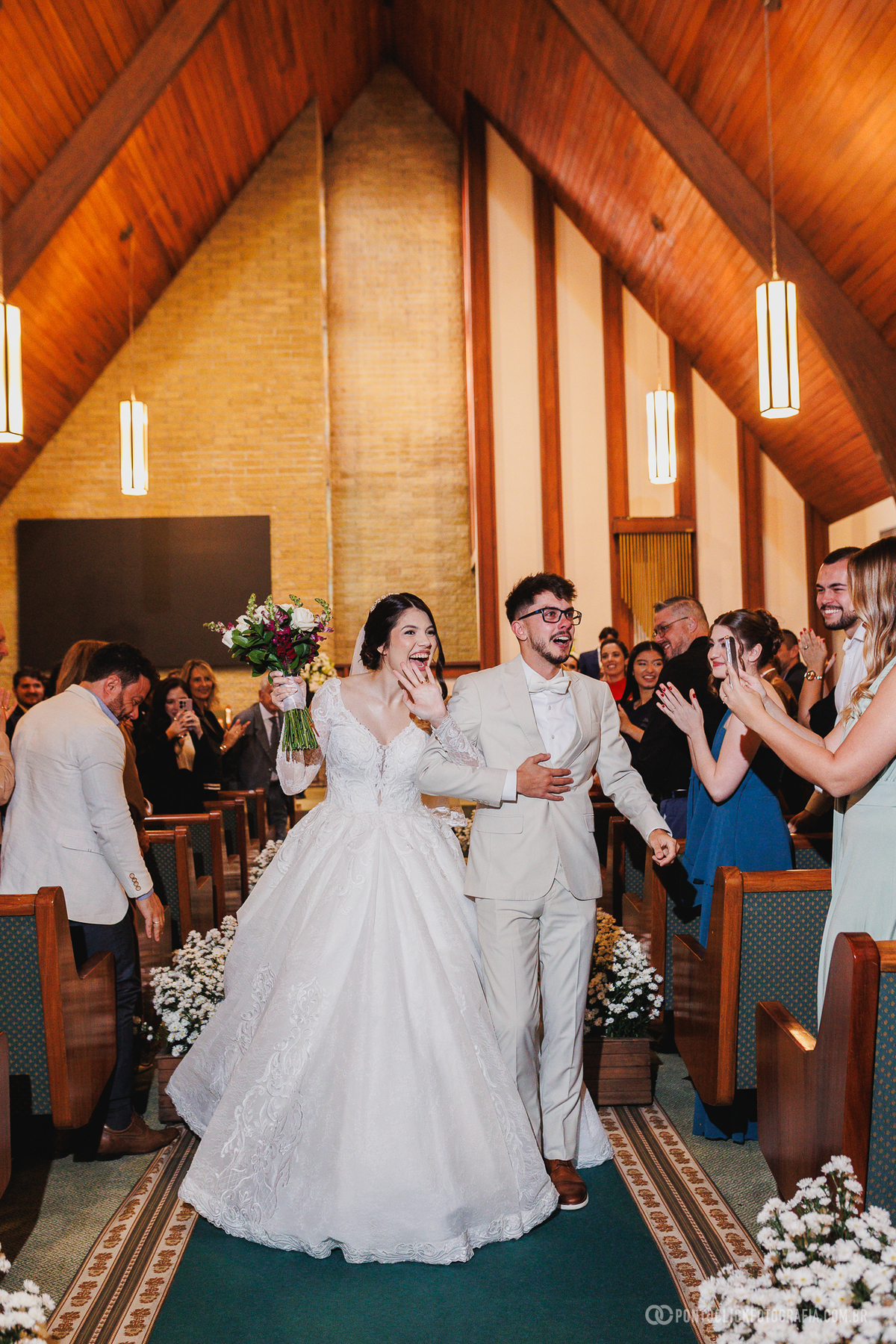 Os noivos percorrem o corredor da igreja após a cerimônia, sendo recebidos com aplausos e sorrisos dos convidados na saída do casamento. Fotógrafo de casamento Pontoclick Fotografia.