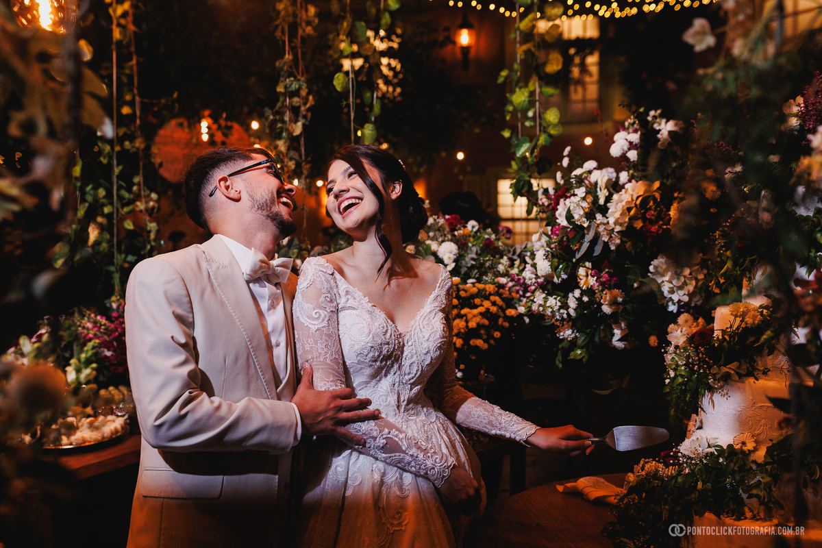 O casal celebra o corte do bolo cercado pela decoração da recepção, registrando um dos momentos mais tradicionais da festa de casamento. Fotógrafo de casamento Pontoclick Fotografia.