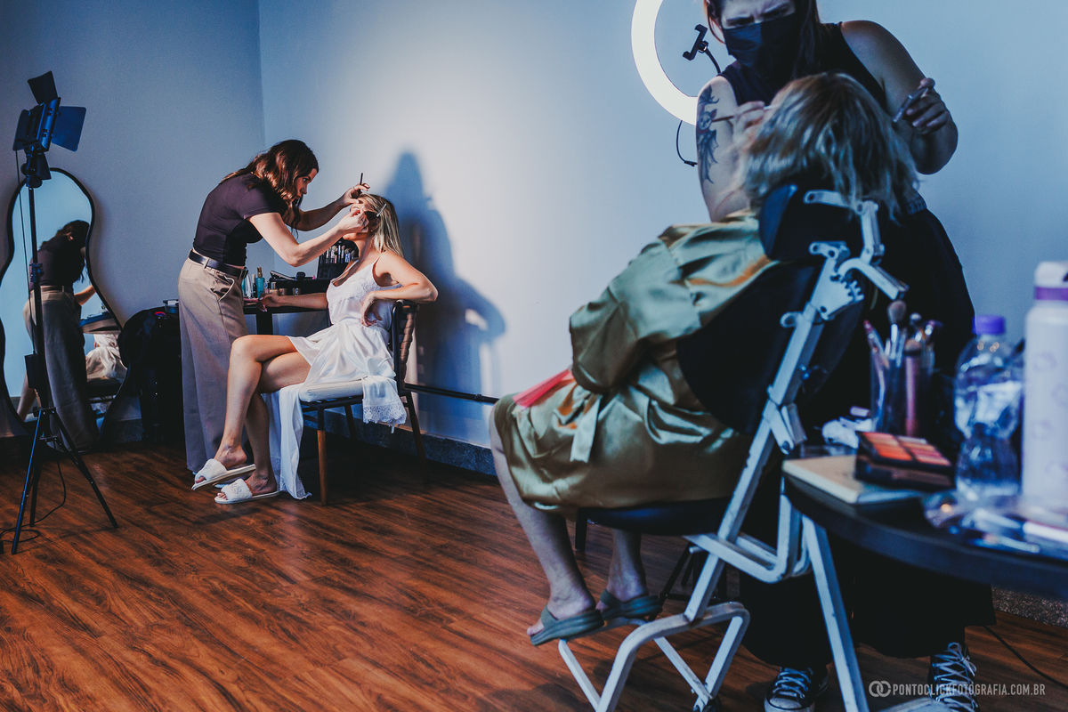 A noiva recebe retoques de maquiagem enquanto está sentada, em um dos momentos dos preparativos para o casamento. Fotógrafo de casamento Pontoclick Fotografia.