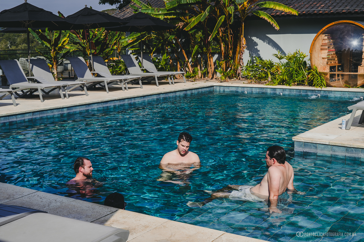 Três homens conversam dentro da piscina, aproveitando um momento de descontração no dia do casamento. Fotógrafo de casamento Pontoclick Fotografia.